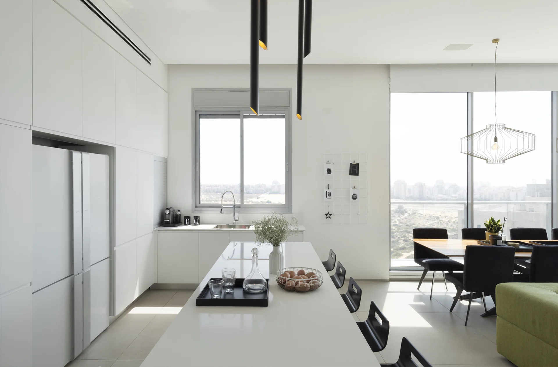 A minimalist white kitchen features handleless integrated cabinets, a long white island with black stools, and black tubular pendant lights.
