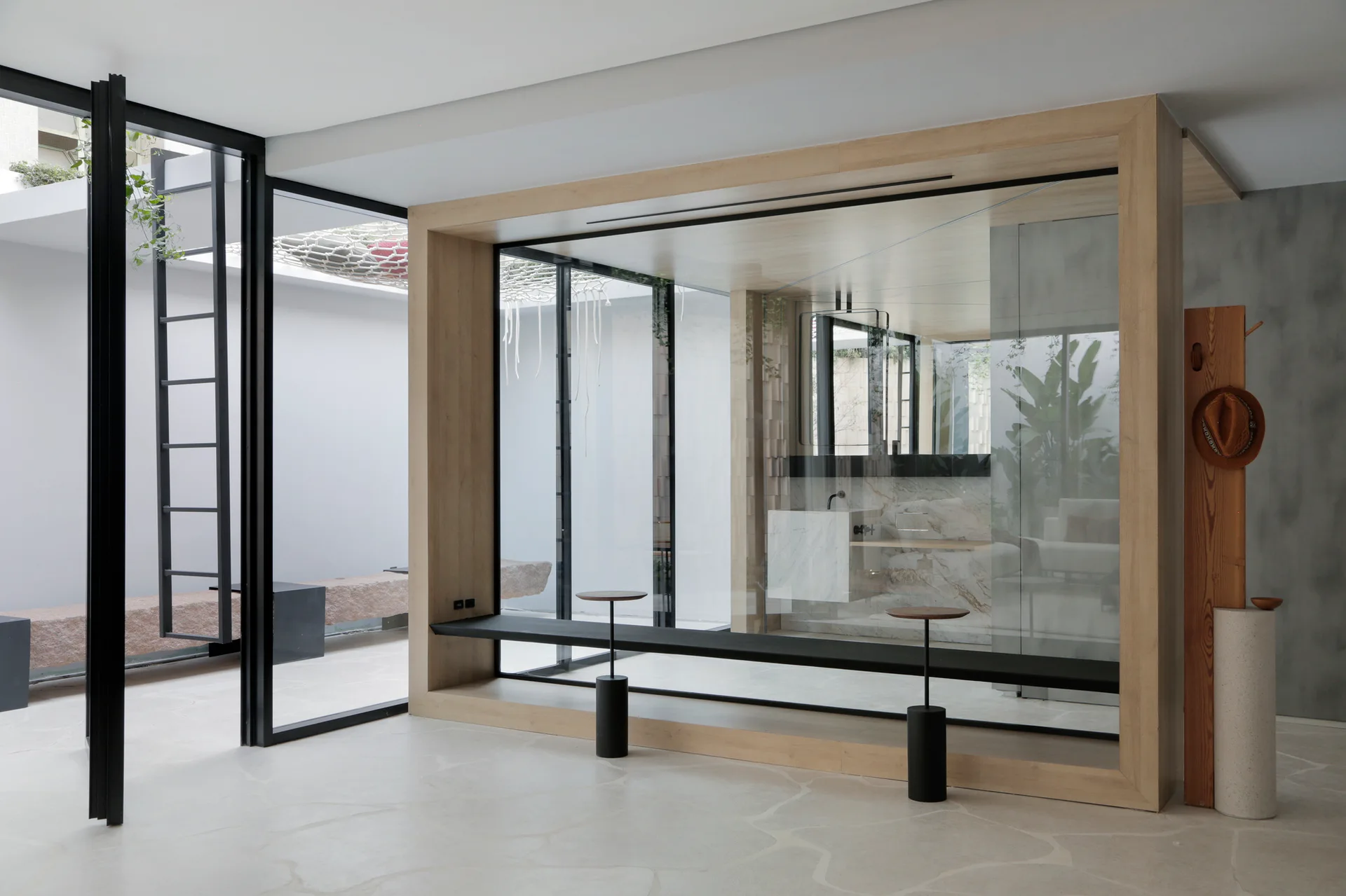 A minimalist glass bathroom enclosure framed in light wood features a marble vanity, situated on organic stone tile flooring near a courtyard.