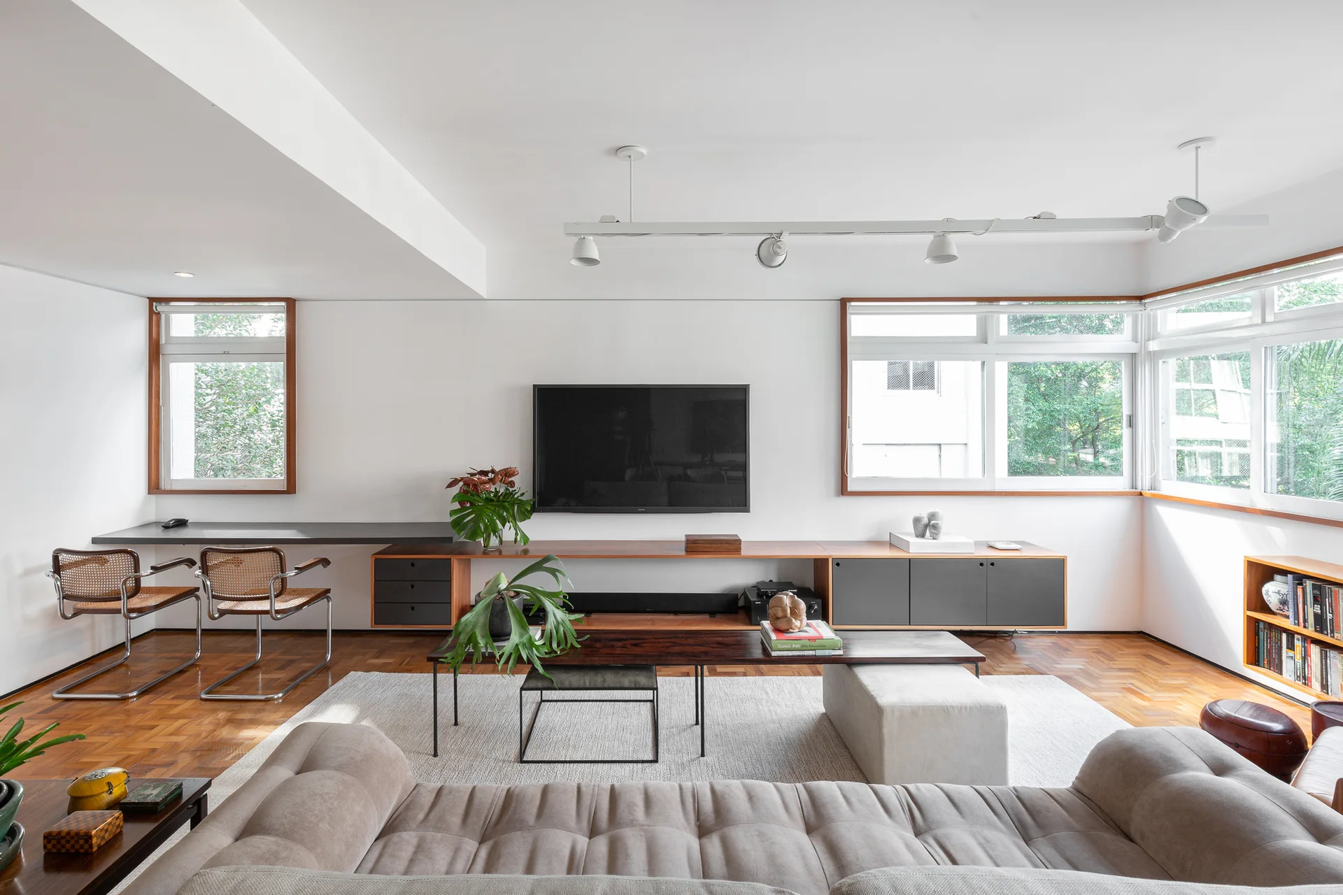A spacious living room featuring a tufted beige sofa, a low-profile wood media console with a wall-mounted TV, and classic parquet wood flooring.
