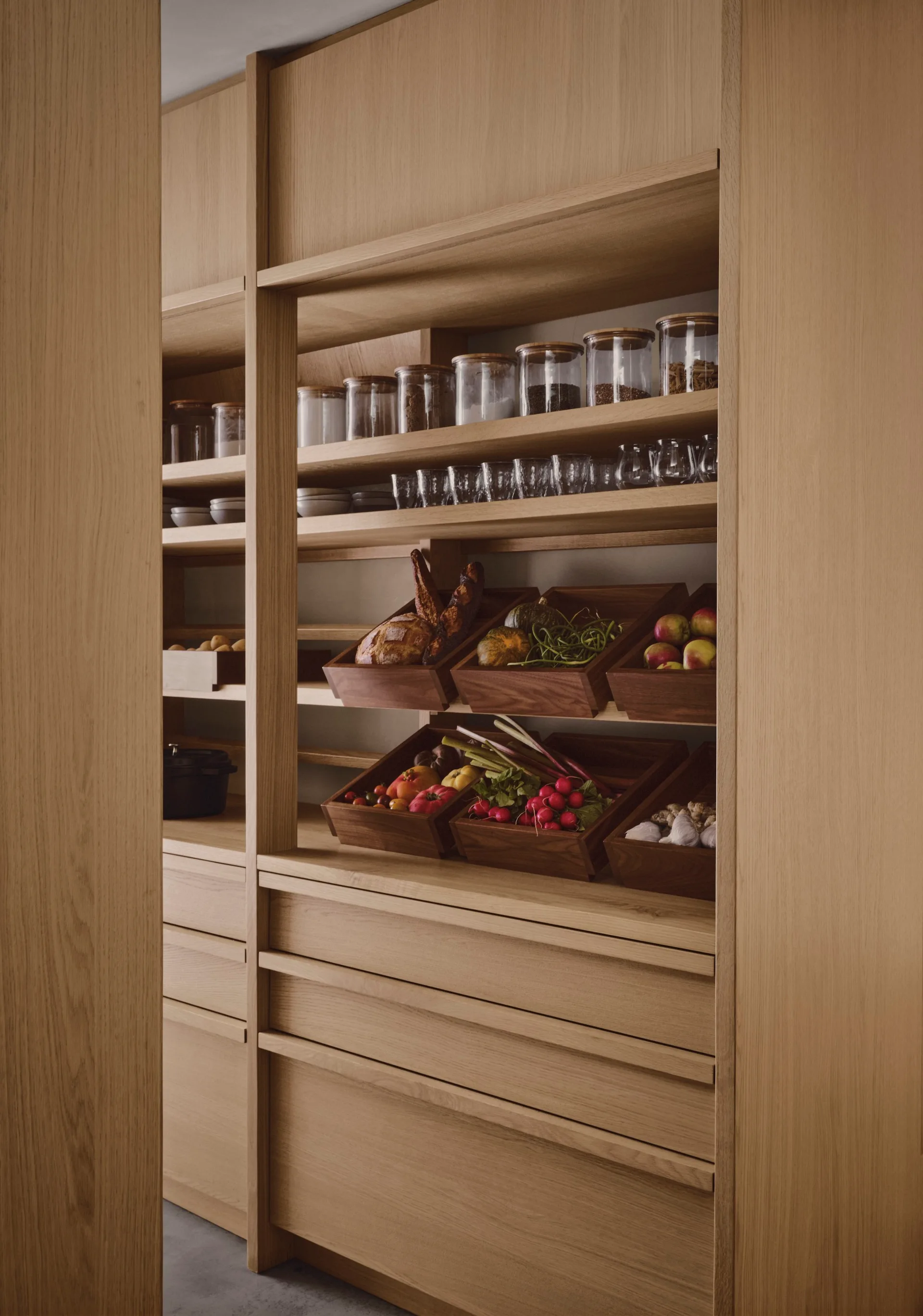 A built-in light wood pantry cabinet features integrated shelves holding glass jars, glassware, and dark wood crates filled with fresh vegetables and fruits.