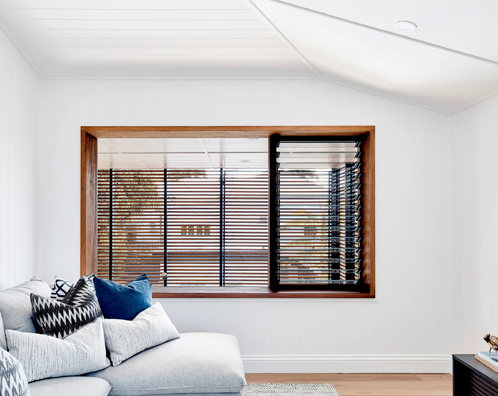 A light gray sectional sofa sits against white vertically paneled walls next to a large window featuring a natural wood frame and slatted blinds.