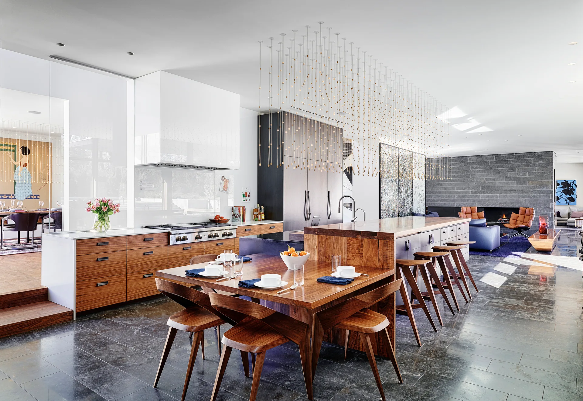 A modern kitchen features a large wood island with an integrated dining table, matching stools, dark stone floors, and a cascading gold ceiling fixture.
