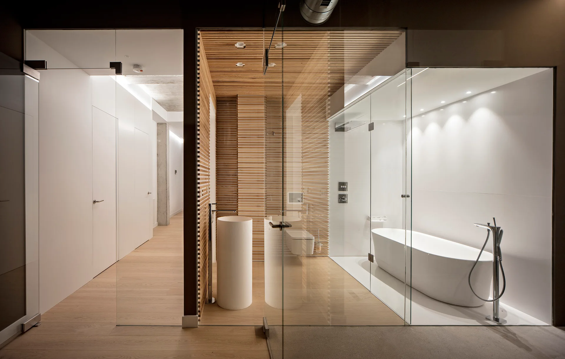 A modern minimalist bathroom features a freestanding bathtub and a cylindrical pedestal sink enclosed by clear glass partitions and light wood slat walls.