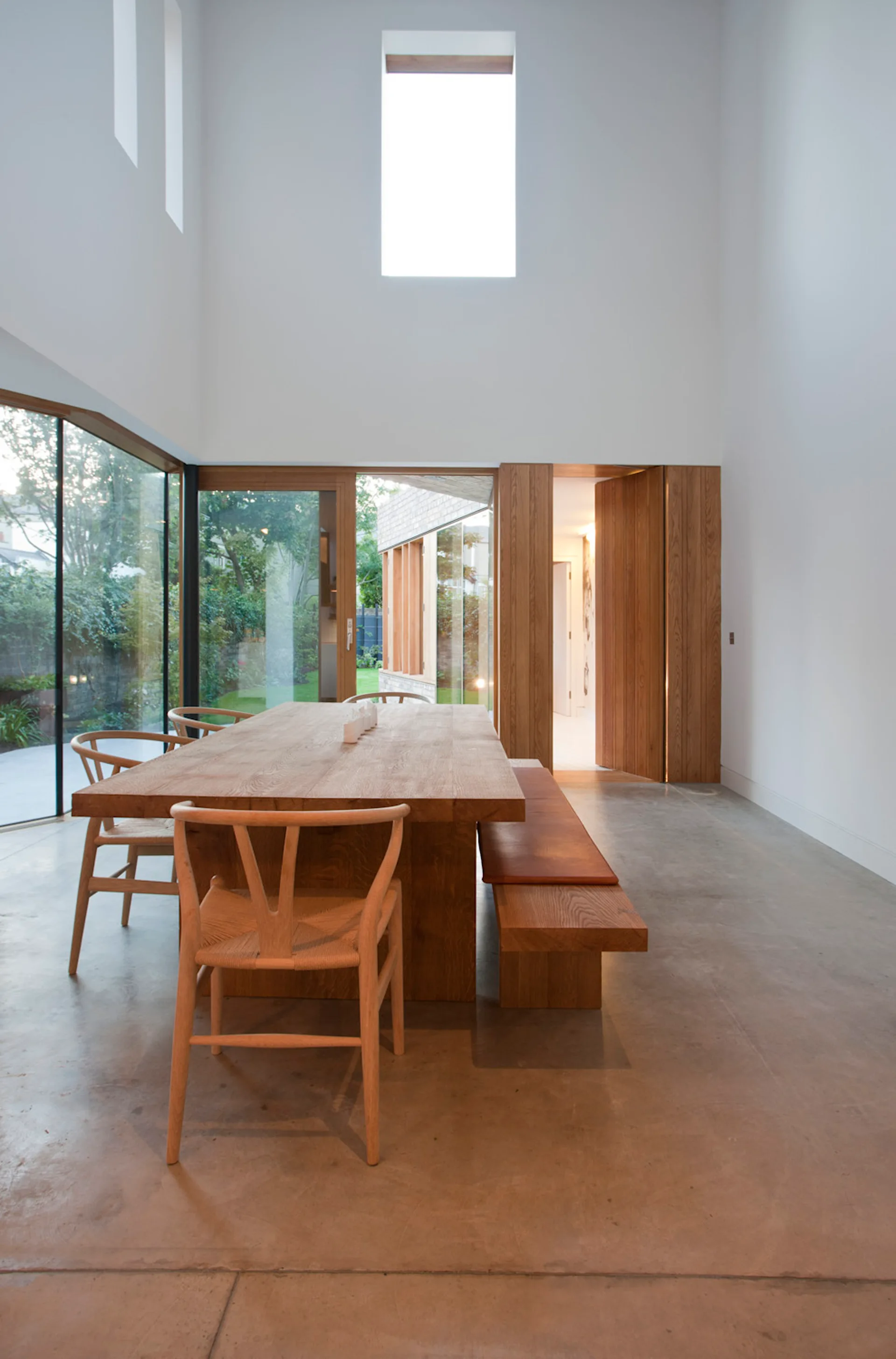 A minimalist dining room features a large oak table, wooden wishbone chairs, and a bench on a polished concrete floor next to glass walls.