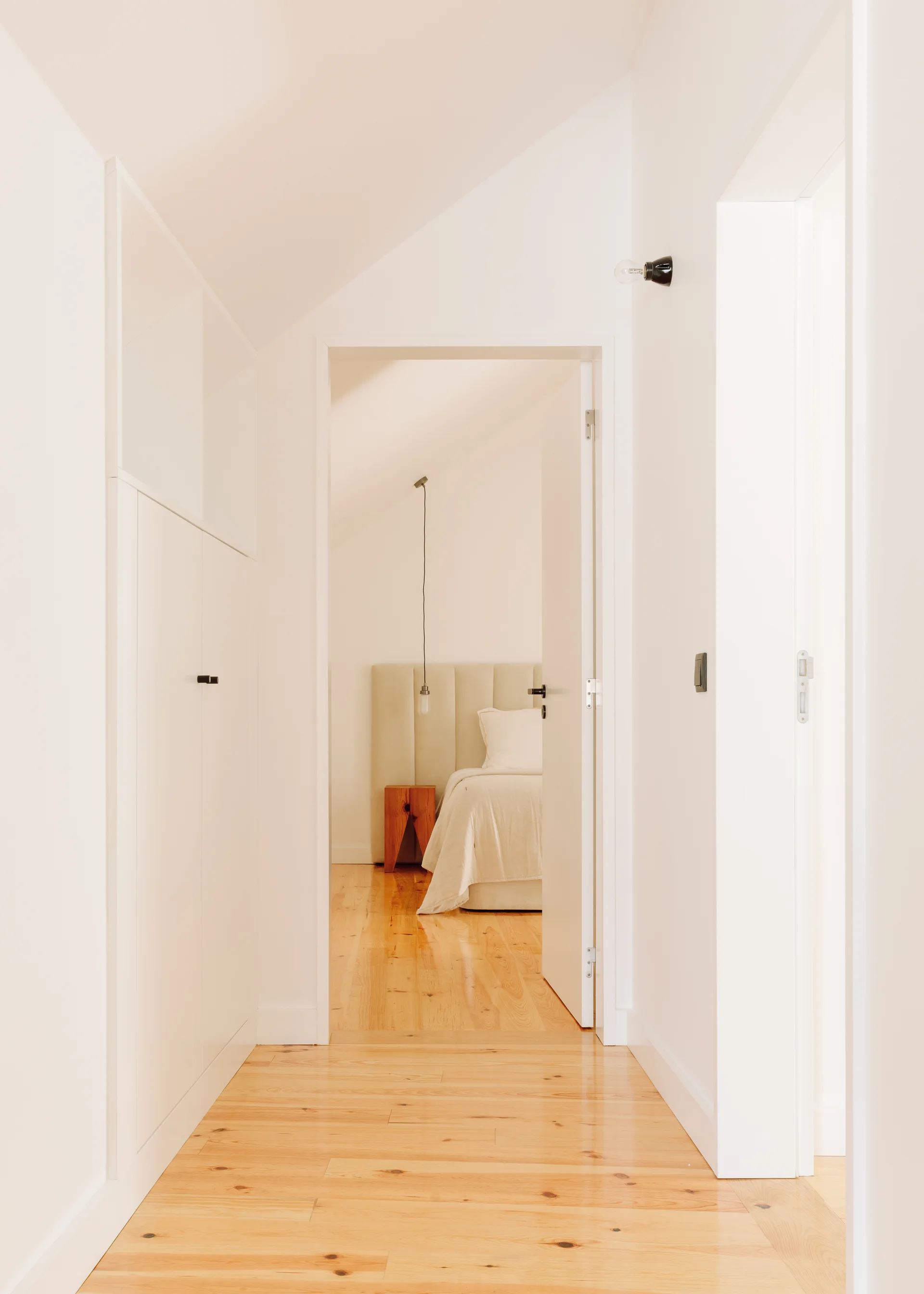 A narrow white hallway with built-in storage cabinets and polished wood floors leads into a bright bedroom with a bed and wood nightstand.