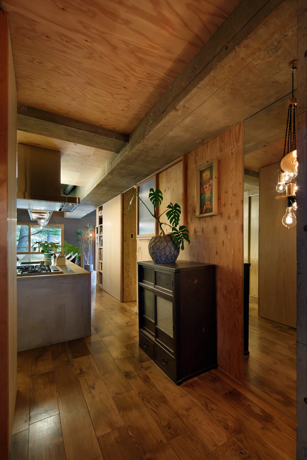 A modern interior corridor showcases warm plywood walls, an exposed concrete beam, a dark wood chest, and a concrete kitchen island in the background.