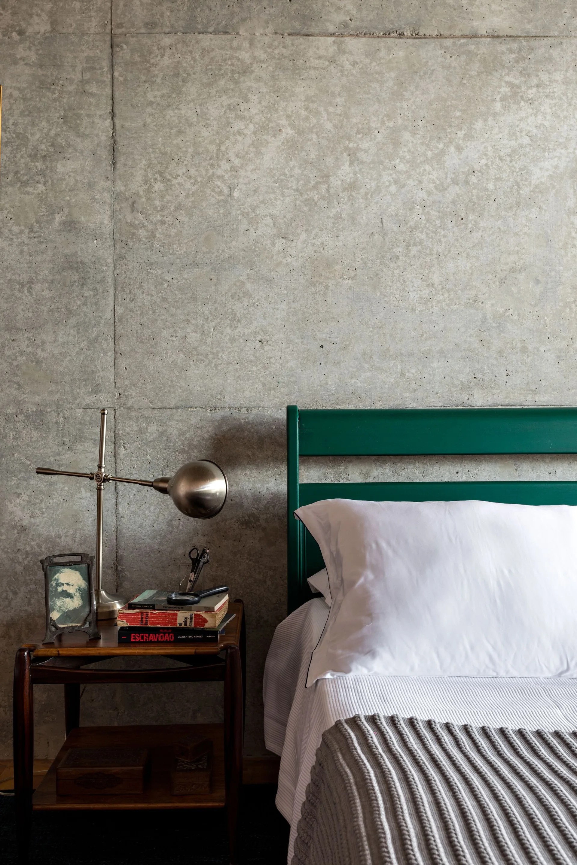A green wooden headboard sits against a gray exposed concrete wall next to a wooden nightstand with a metal lamp and books.