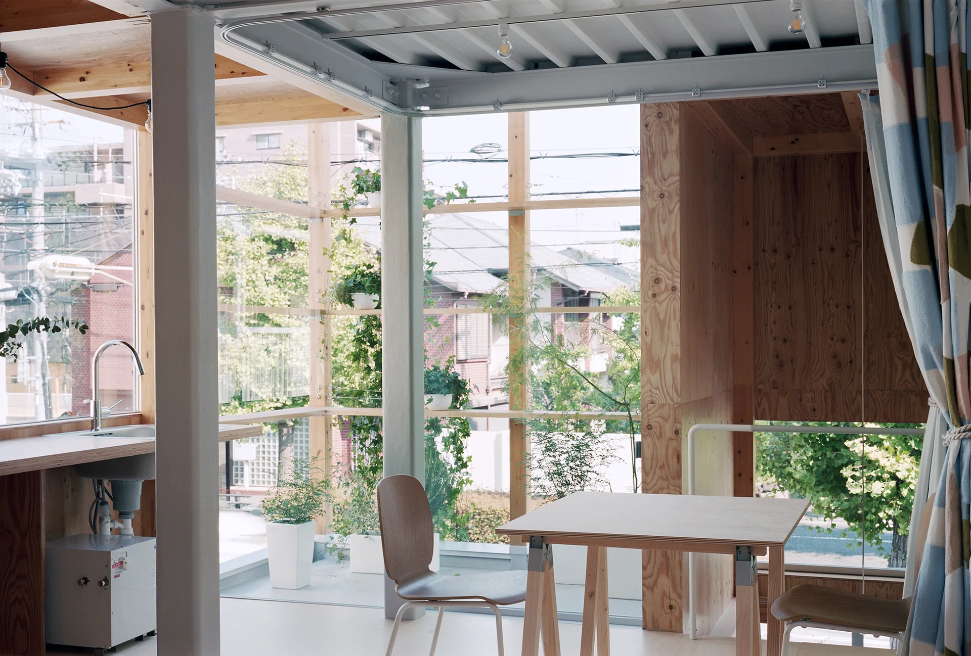 A minimalist dining room featuring a wooden table, simple chairs, exposed plywood walls, and large glass windows overlooking residential buildings.