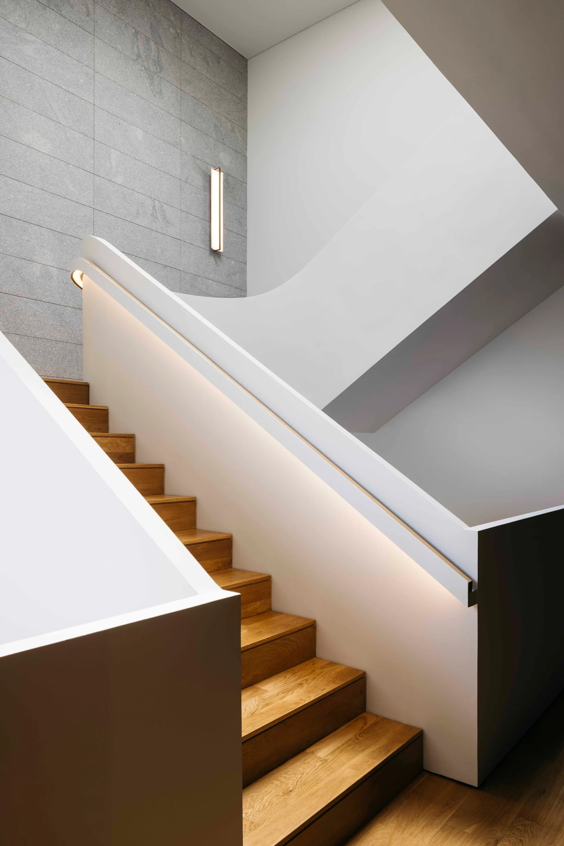 A modern wooden staircase features warm oak treads, a white curved railing with integrated LED lighting, and a gray stone tiled wall.