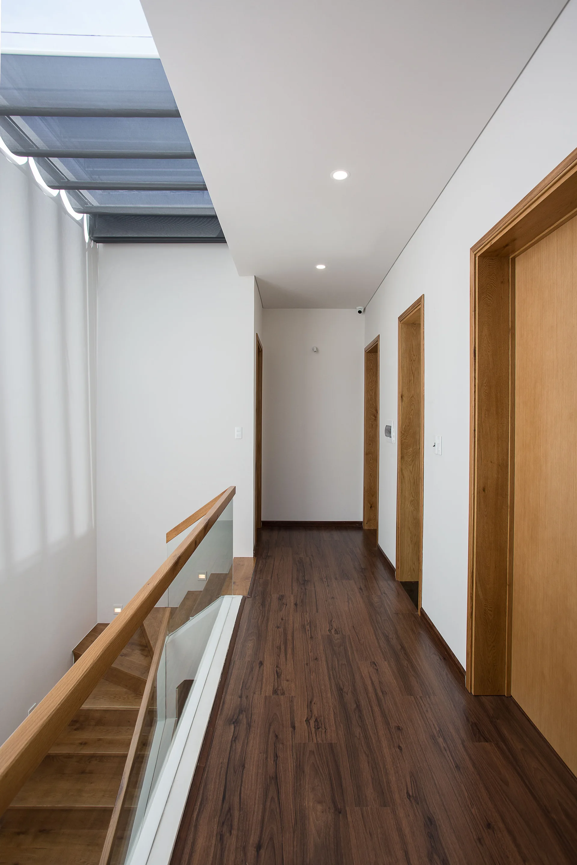 A modern hallway features dark wood flooring, white walls, light wood doors, and a staircase with a glass railing under a large skylight.