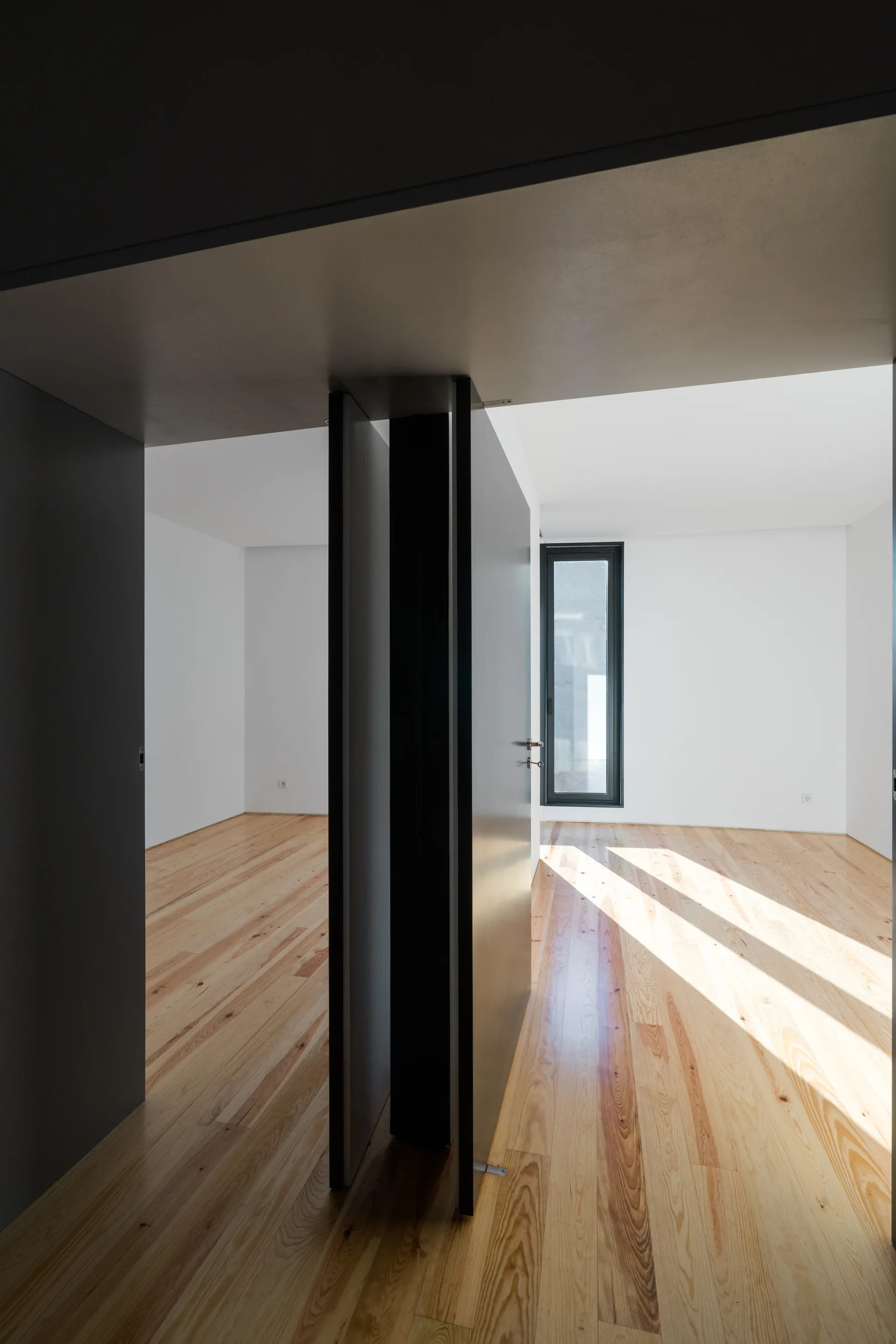 An open dark grey door stands between a minimalist hallway and a bright room featuring light wood floors and a narrow vertical window.