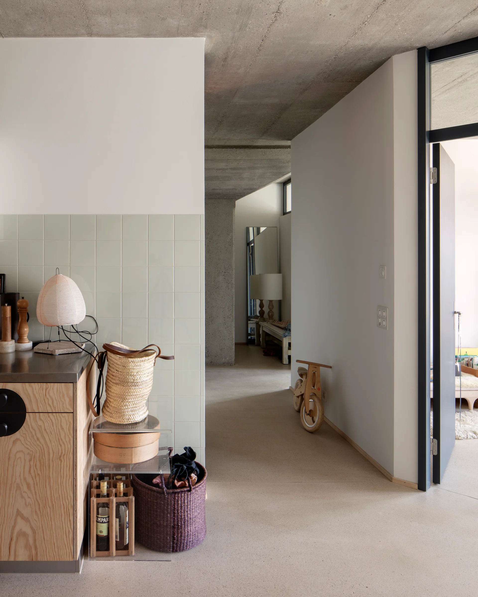 A modern kitchen entryway features plywood cabinetry, a pale green tile backsplash, and an exposed raw concrete ceiling above a polished floor.