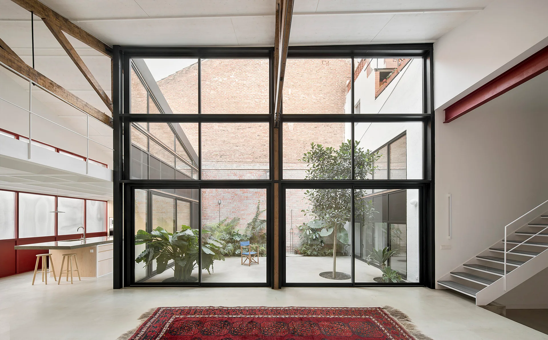 A modern double-height living room features a massive black-framed glass wall looking out onto a central courtyard with plants and a tree.