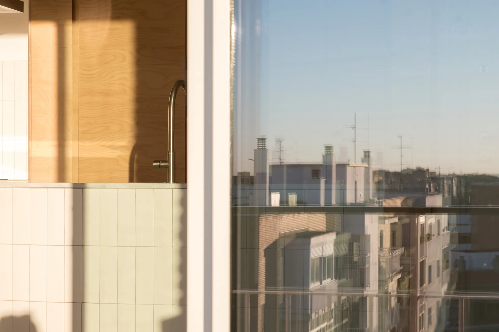 A close-up of a sleek metal kitchen faucet against a cream vertical tiled backsplash, next to a window reflecting city buildings.