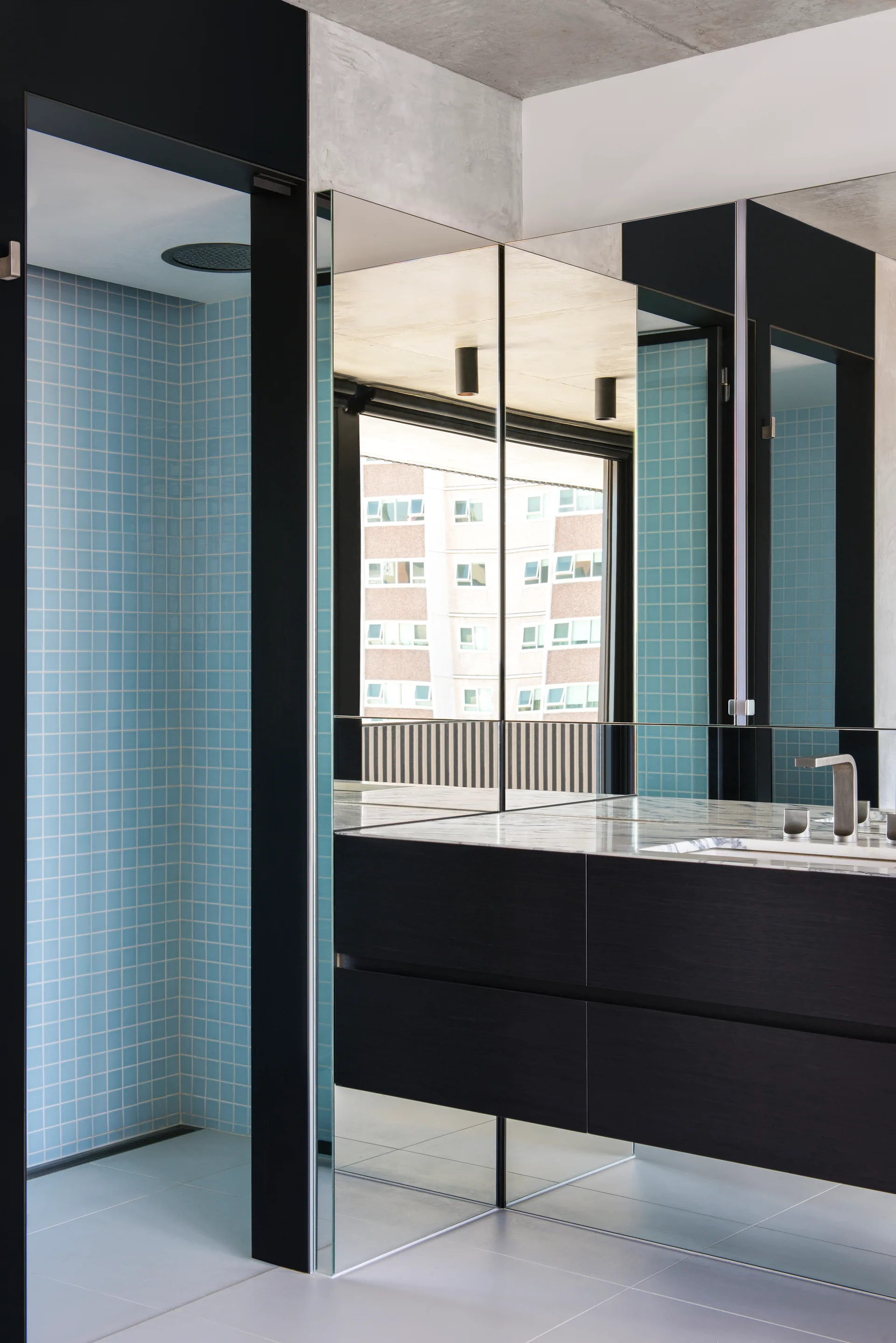 A modern bathroom features a dark floating vanity under a mirrored wall, adjacent to a walk-in shower with light blue grid tiles.