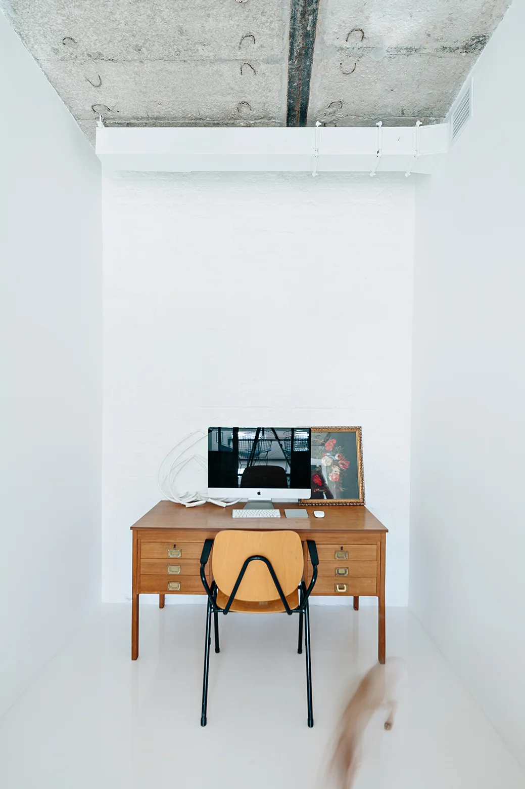 A mid-century wooden desk and chair sit in a stark white alcove office beneath an exposed concrete ceiling.
