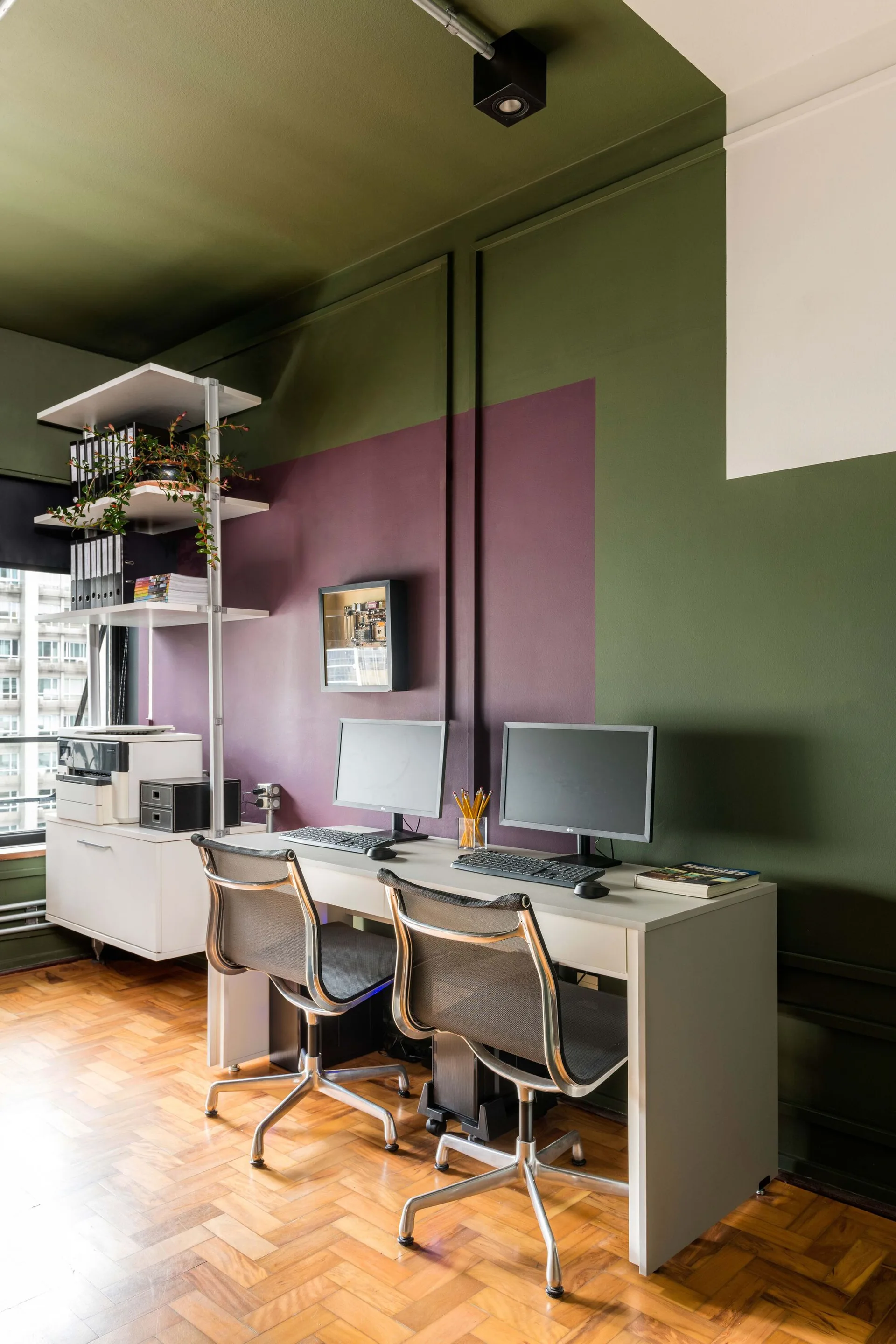 A home office features a dual workstation desk with two monitors and mesh chairs against a painted green and purple color-blocked wall.