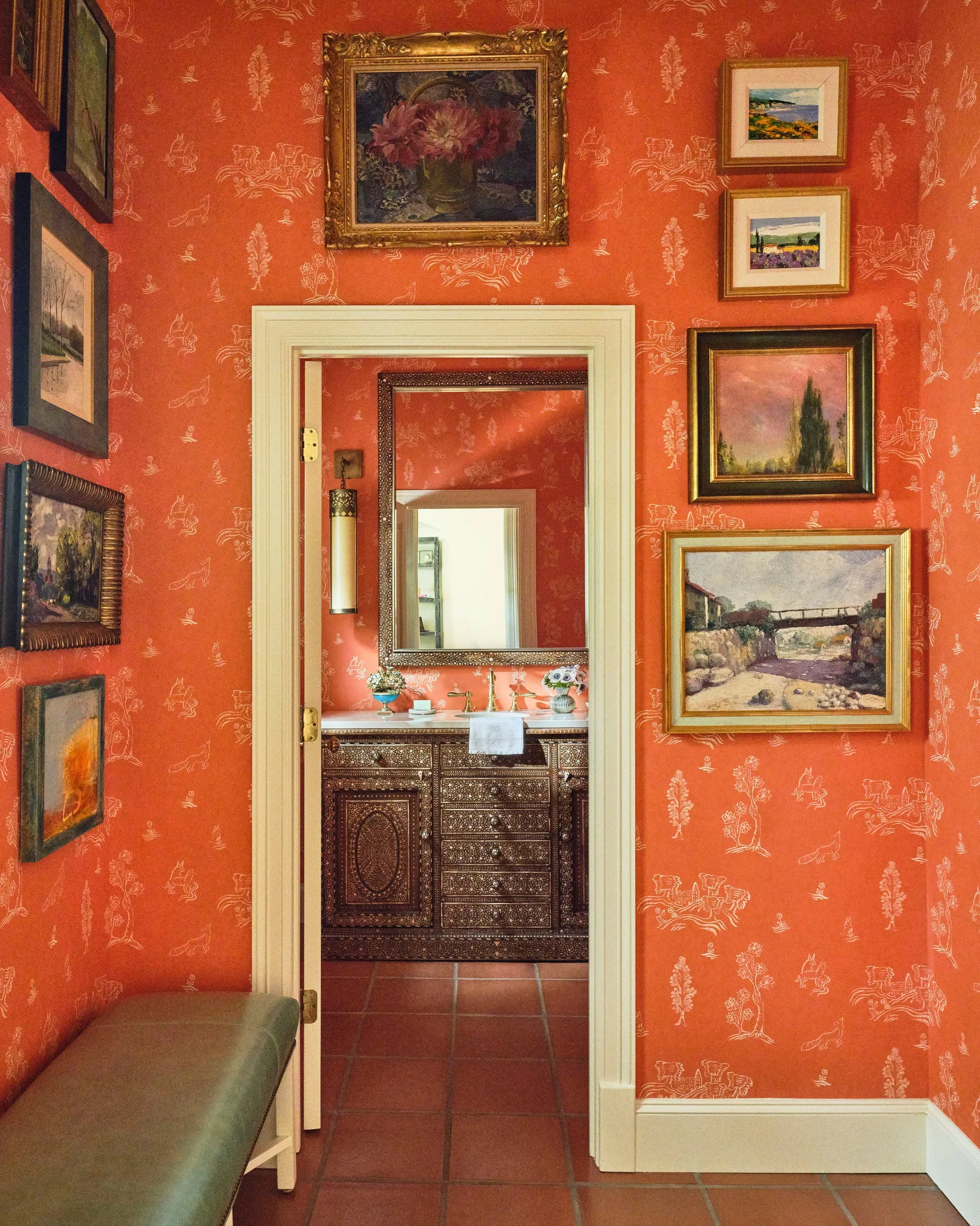 An entryway featuring orange toile wallpaper and a gallery wall of framed paintings leads to a powder room with an ornate wood vanity.