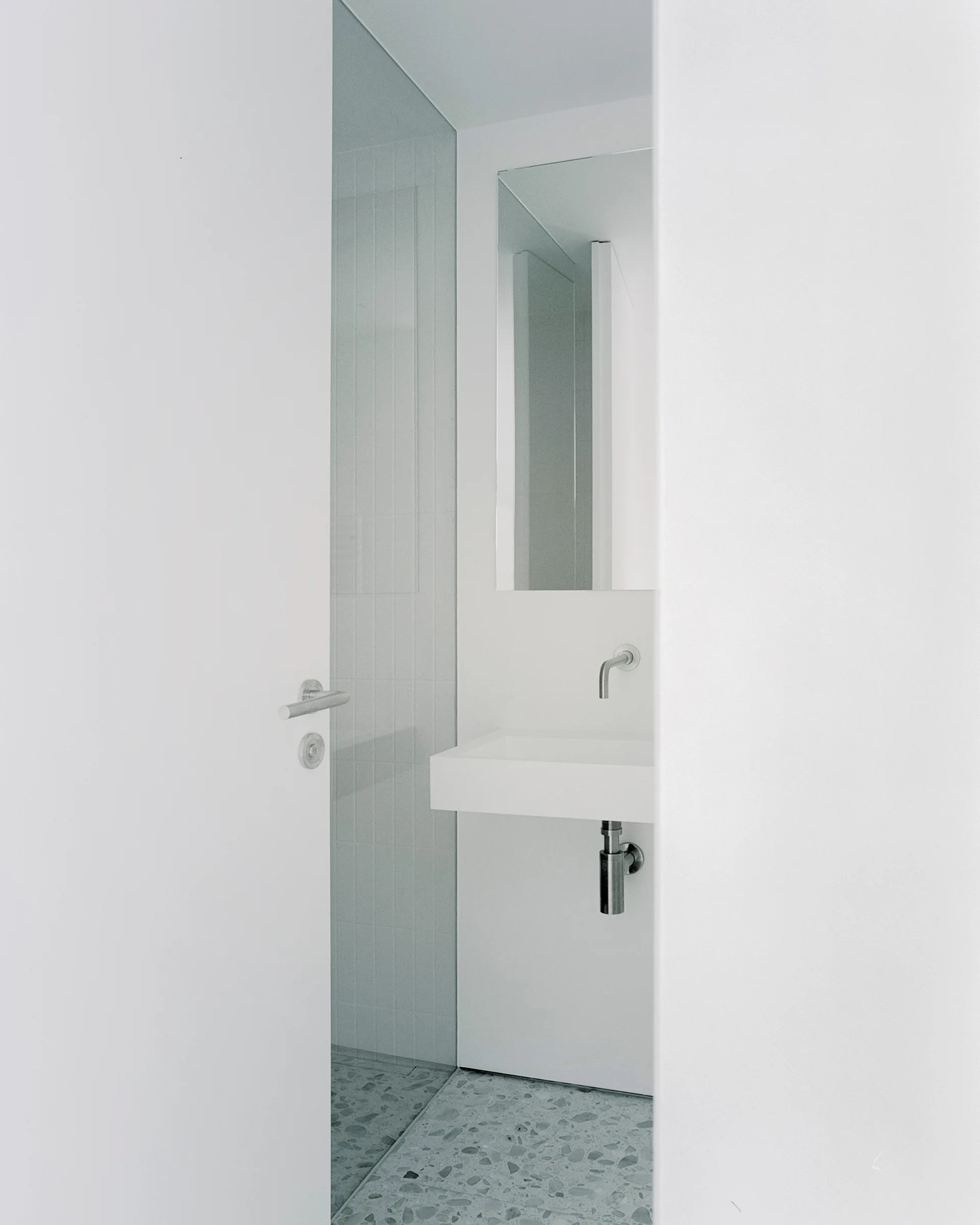 A minimalist white bathroom features a wall-mounted rectangular sink, a recessed mirror, and grey terrazzo flooring viewed through an open doorway.