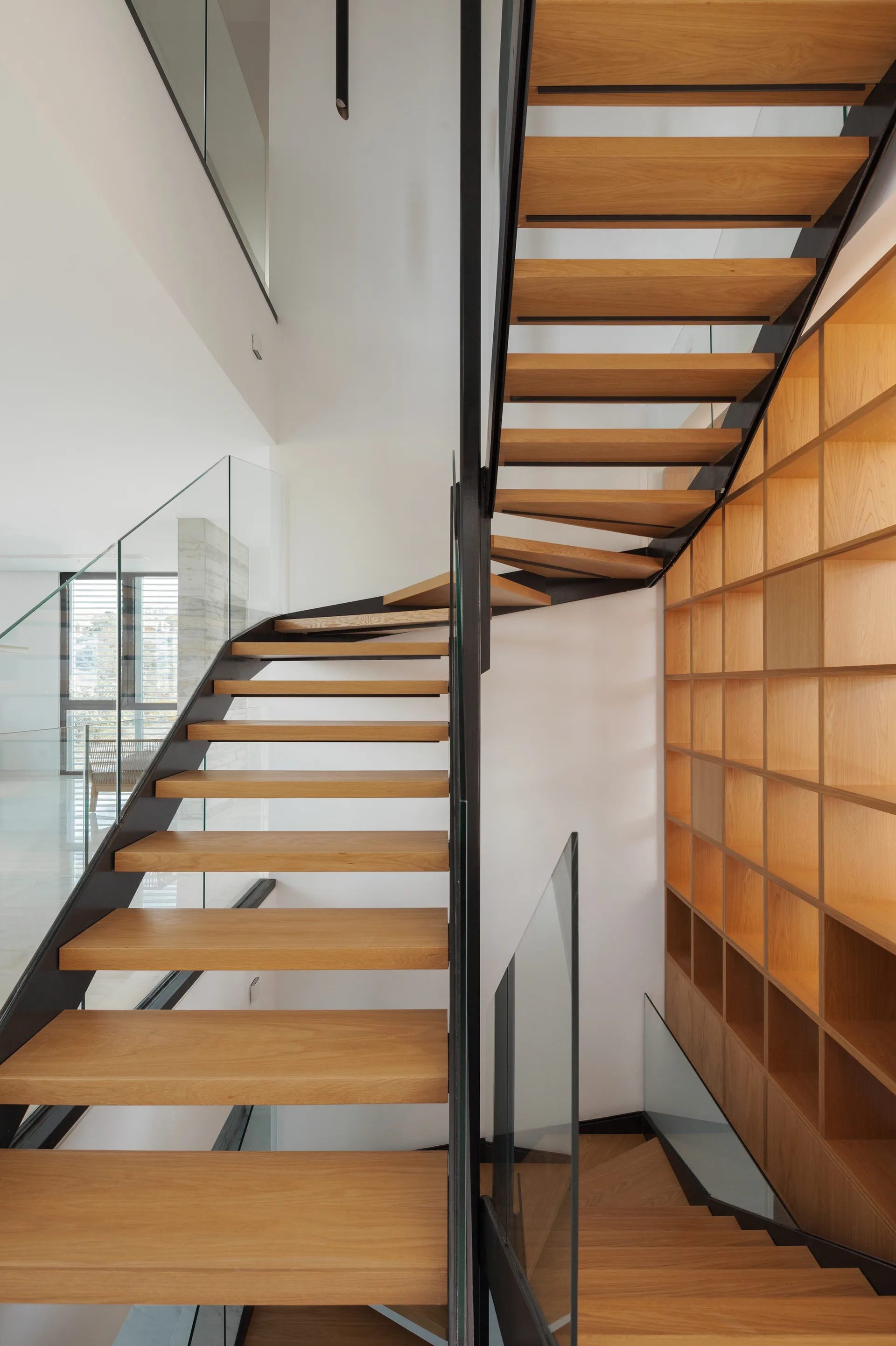 A modern open-tread wooden staircase with a black metal frame and glass railings stands next to a full-height wooden grid bookshelf.