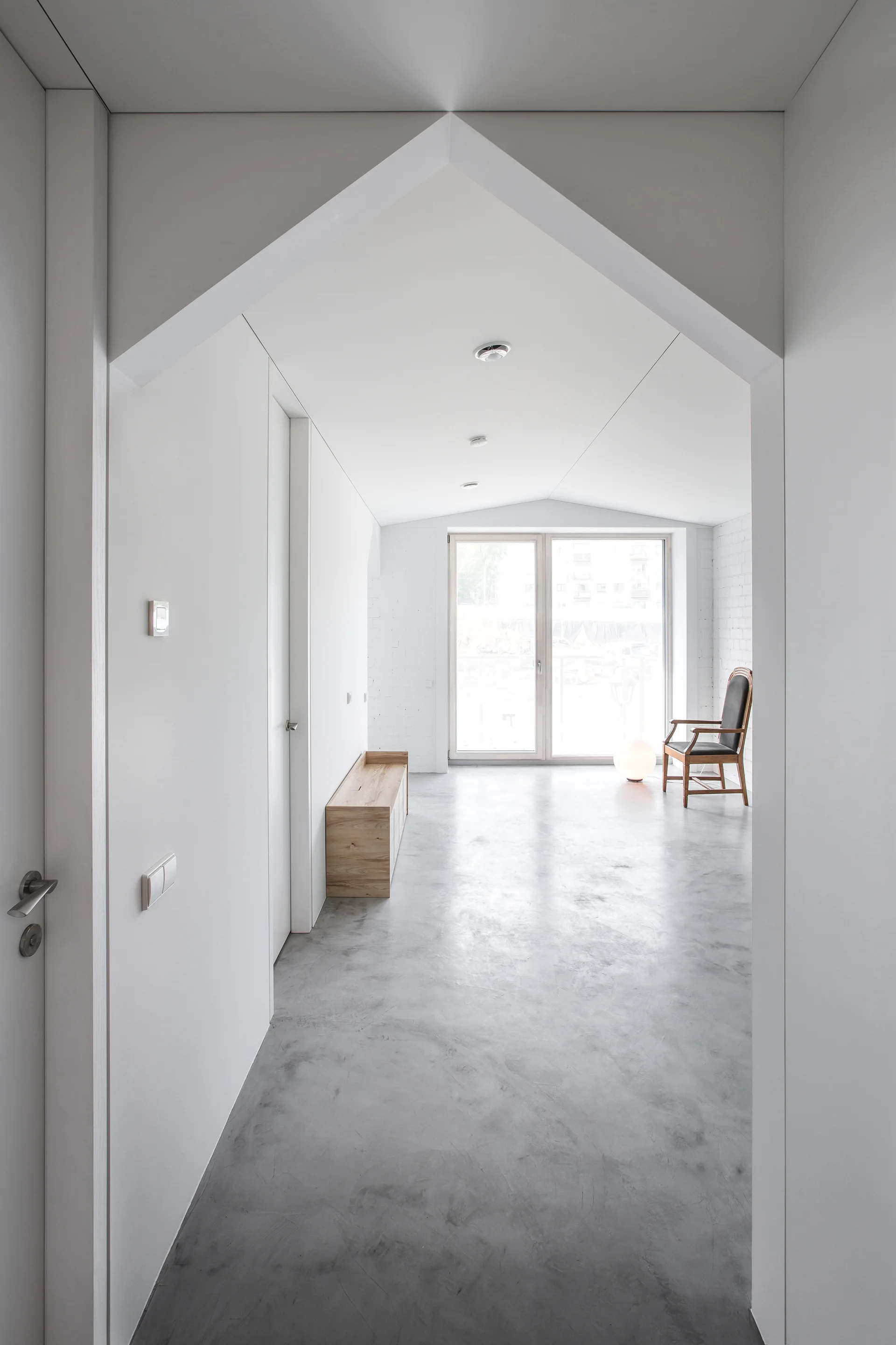 A minimalist hallway with a peaked archway features polished grey concrete floors, a low wooden bench, and a large window at the far end.