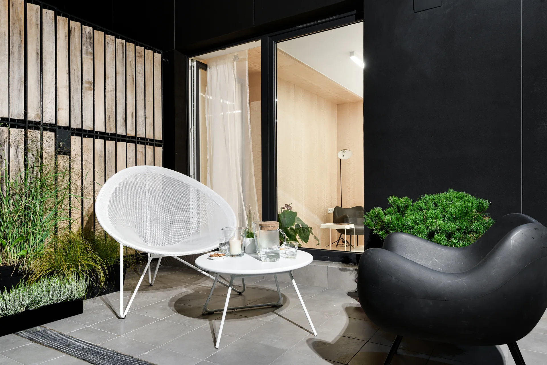 A modern outdoor patio features a white mesh chair, a sculptural black chair, and a small round table on a grey tiled floor next to a wood privacy screen.