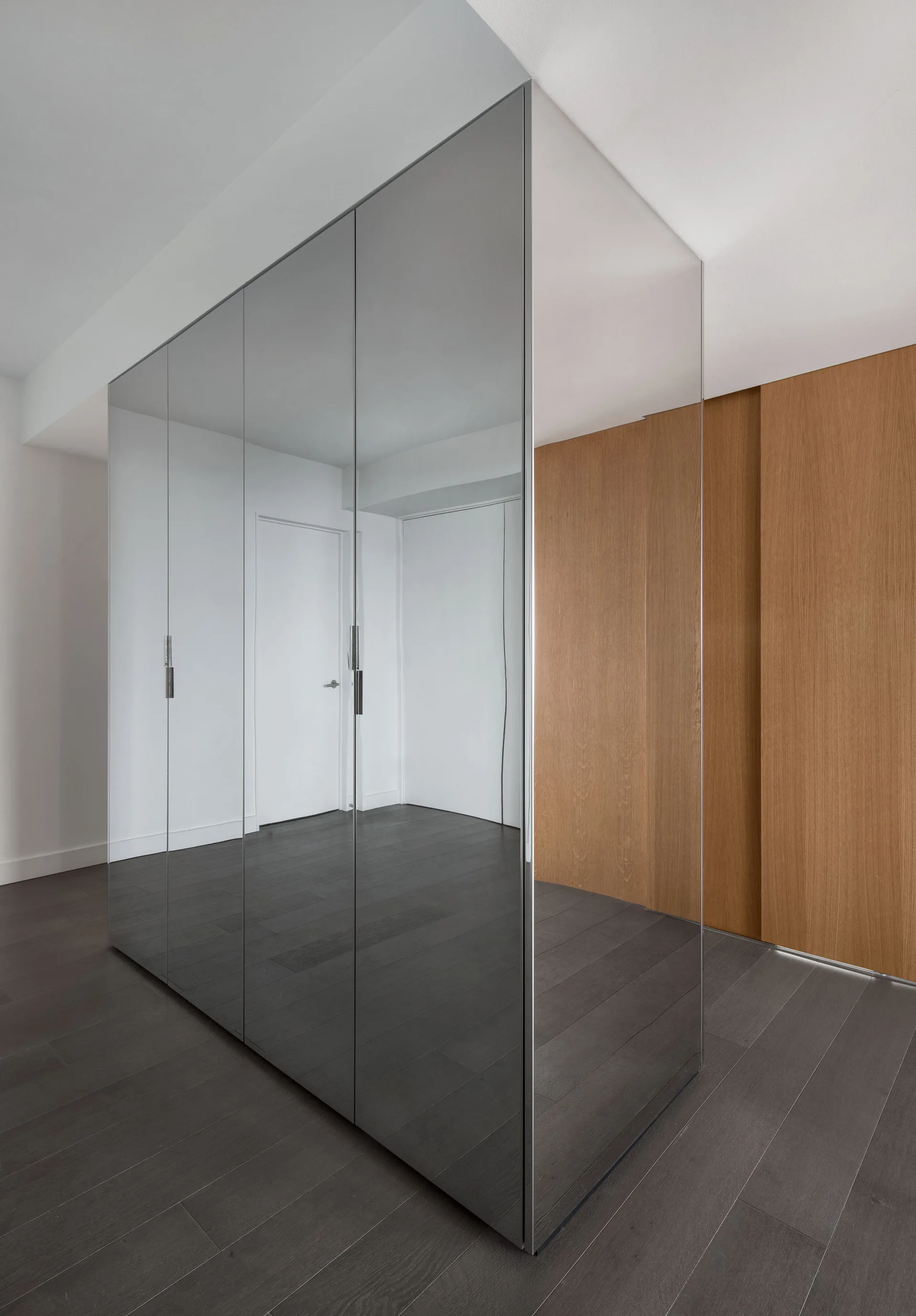 A minimalist hallway features a floor-to-ceiling mirrored wardrobe unit beside a natural wood paneled wall and dark wood flooring.