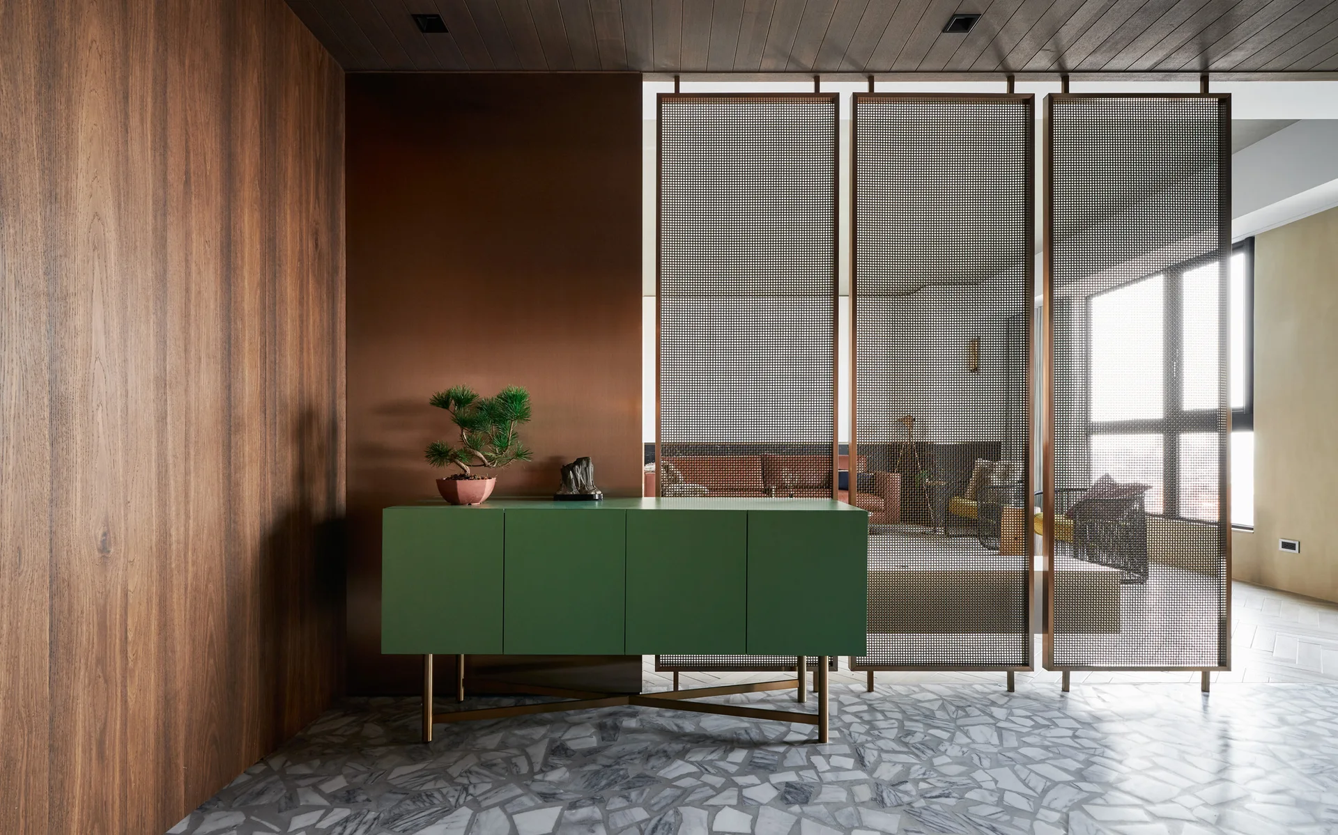 A green console table stands against a metallic wall panel next to bronze mesh room dividers on a grey stone fragment floor.
