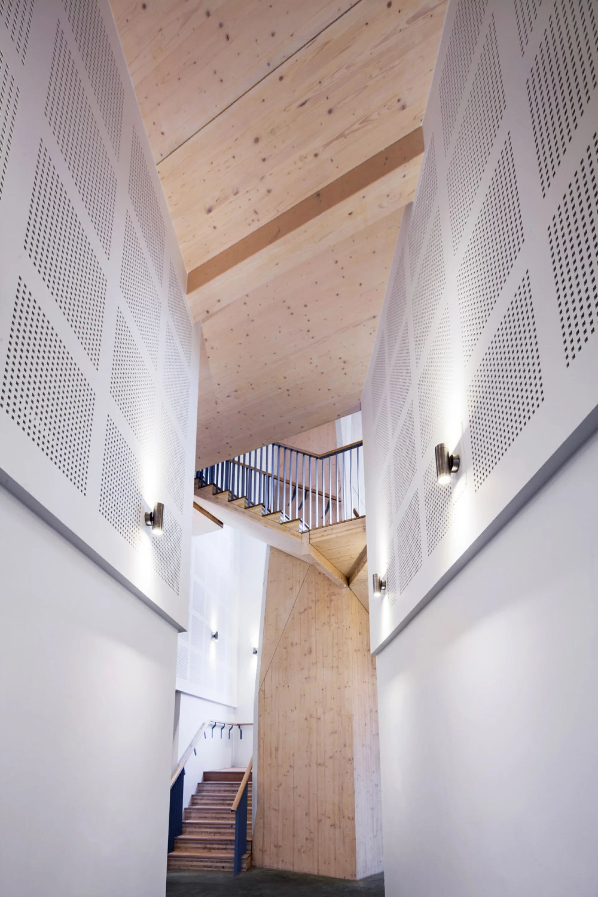 A modern staircase made of light wood with dark railings, framed by white perforated wall panels with small cylindrical sconces.