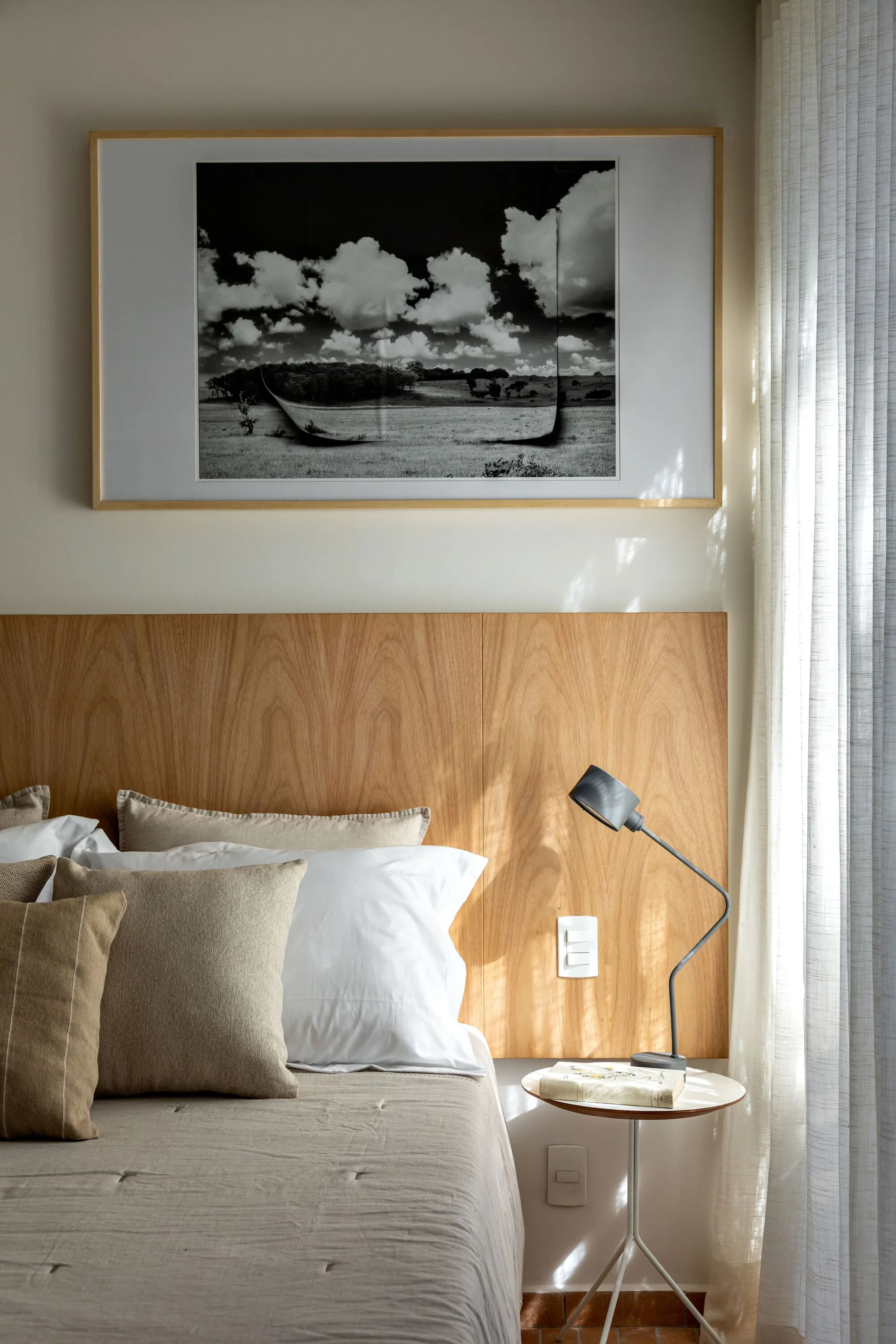 A bedroom with a warm wood panel headboard, neutral-toned linen bedding, a small round side table with a grey metal lamp, and framed black-and-white art.