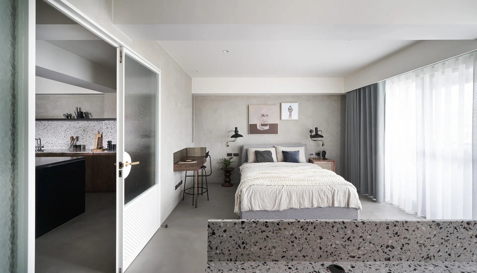 A modern grey bedroom featuring an upholstered bed, a wall-mounted wooden desk, and a white framed fluted glass door leading to a kitchen.