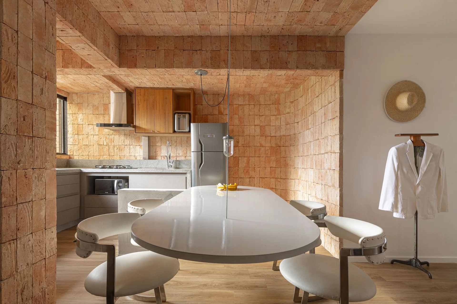 A modern dining space features a glossy white oval table and white chairs beneath a vaulted ceiling and walls made of exposed terracotta brick.