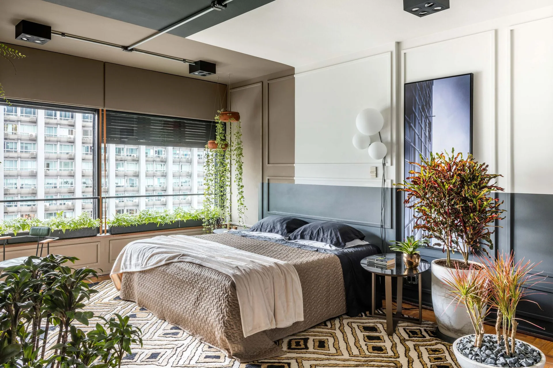 A contemporary bedroom features a bed with dark bedding, a two-tone color-blocked paneled wall, a geometric rug, and numerous potted plants.