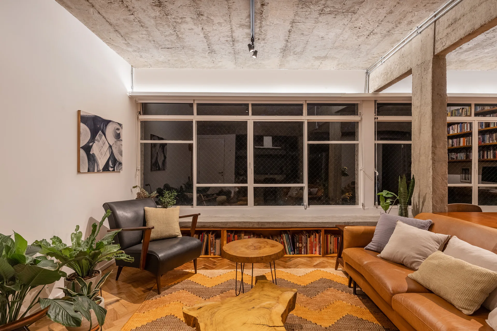 A warm living room features a tan leather sofa, wooden slab coffee tables, and a low bookshelf under a large window with an exposed concrete ceiling.