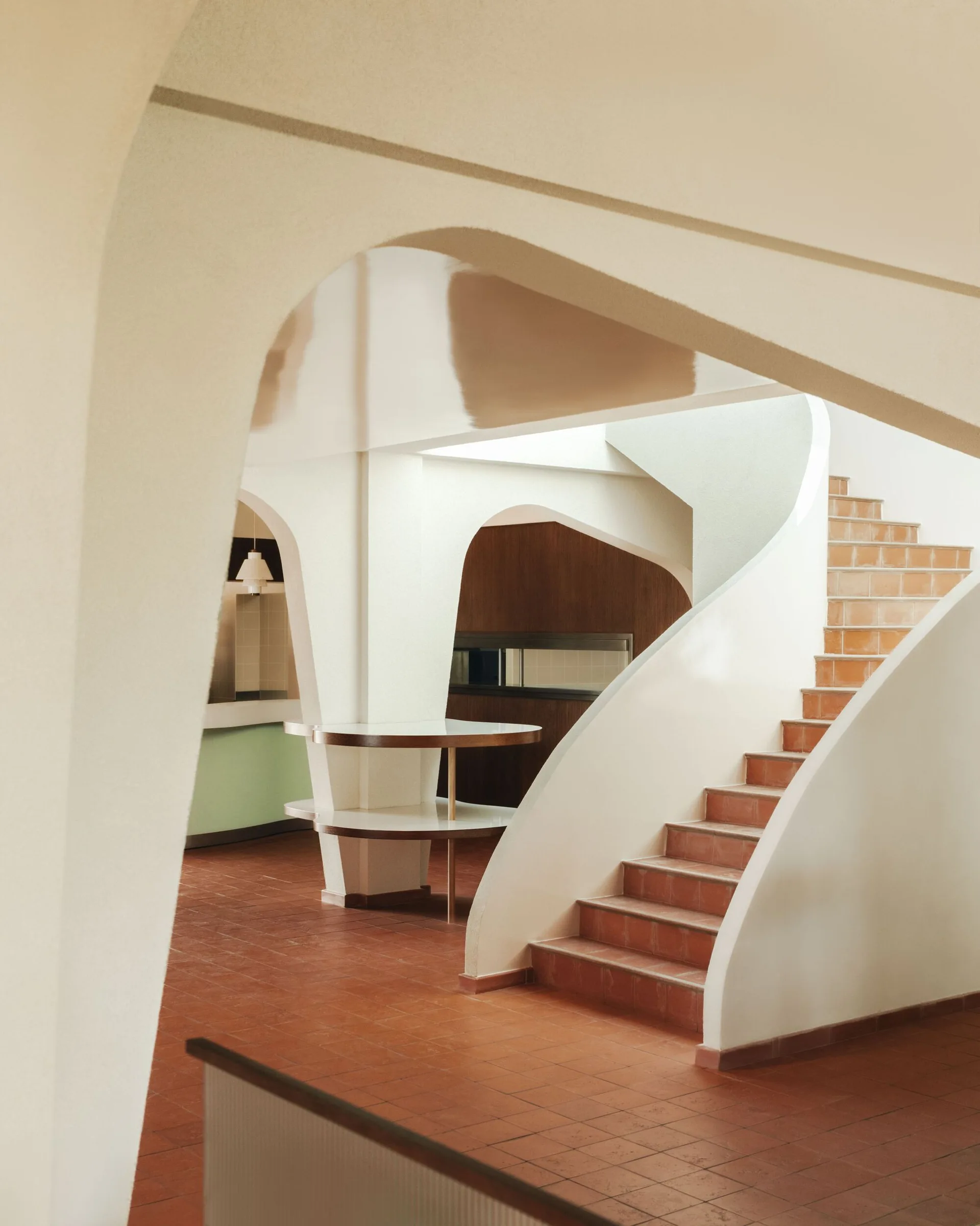 An interior view of a modernist space featuring a curved white plaster staircase, arched openings, and terracotta tiled floors.