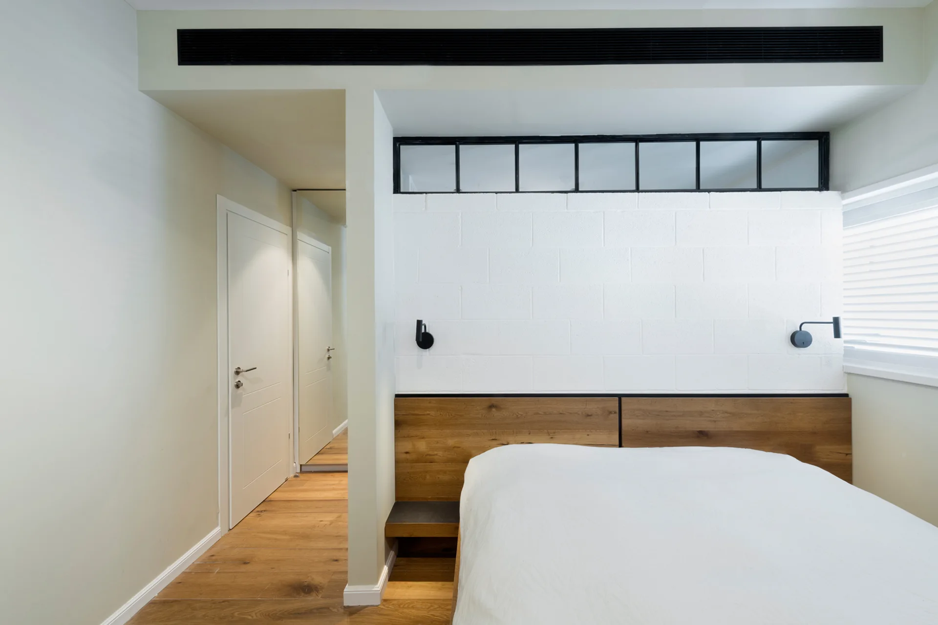 A minimalist bedroom features a bed with a warm wood headboard against a white painted cinder block wall, adjacent to a hallway with wood flooring.