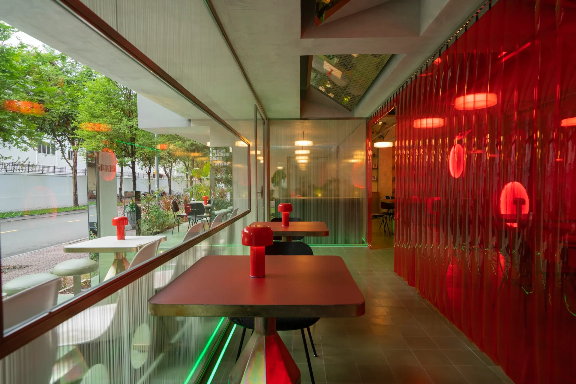 A modern cafe interior features square tables with red mushroom lamps, a red plastic strip curtain, and ribbed glass partition walls next to a window.