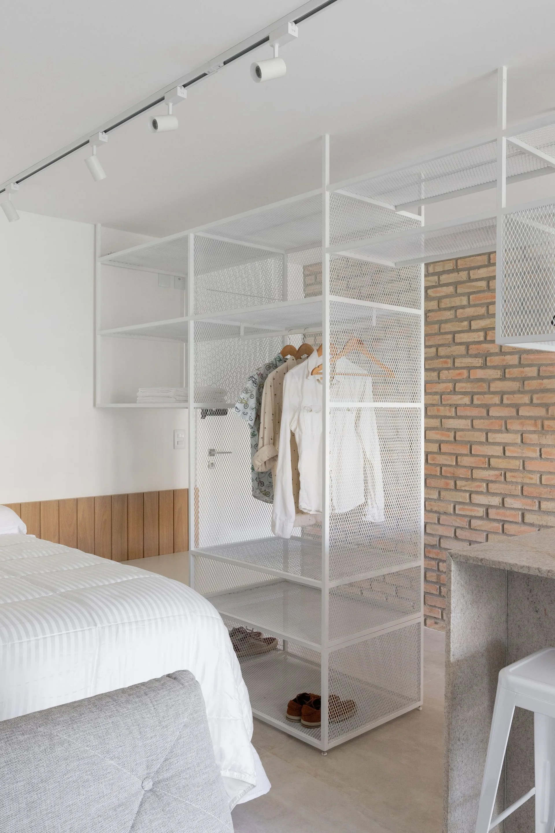 A bedroom features a white metal mesh open shelving closet system alongside a bed with white bedding and an exposed brick wall in the background.