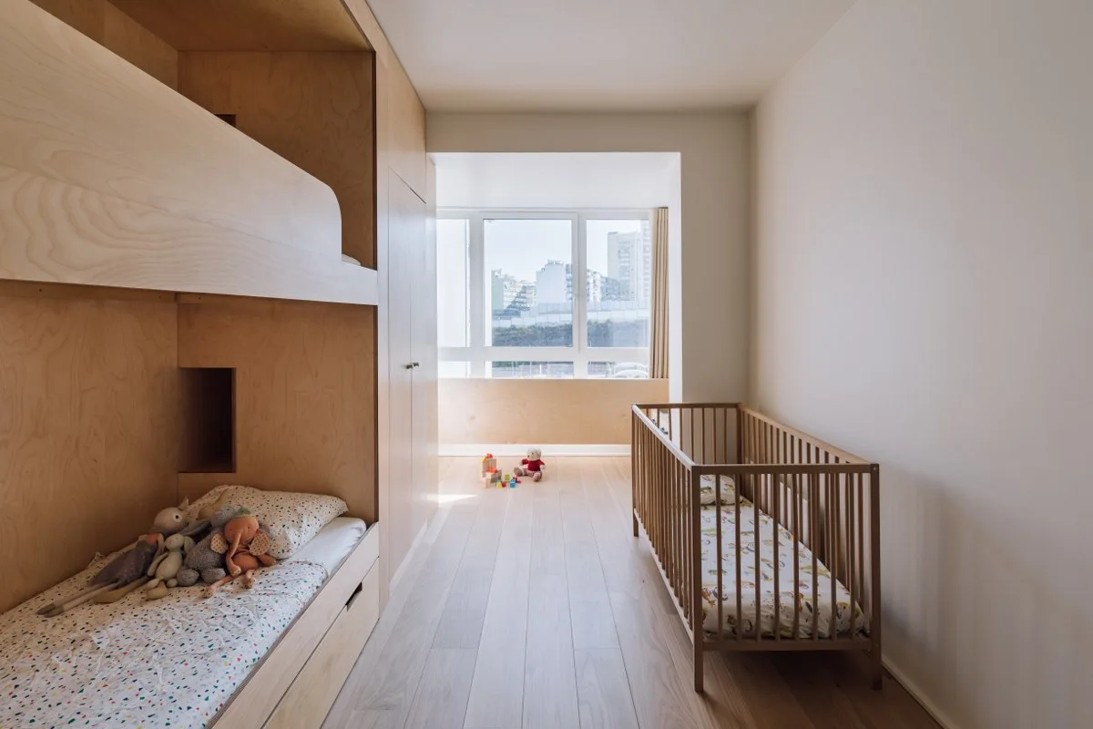 A minimalist children's bedroom featuring a built-in wooden bunk bed, a matching wooden crib, light hardwood floors, and a large window.