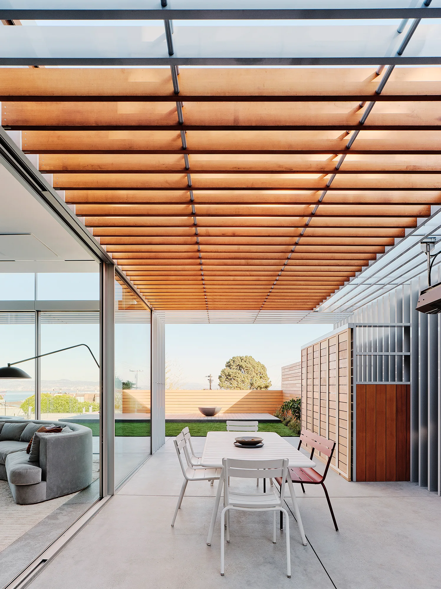 A white modern dining set sits on a concrete patio under a slatted wood pergola next to sliding glass doors.