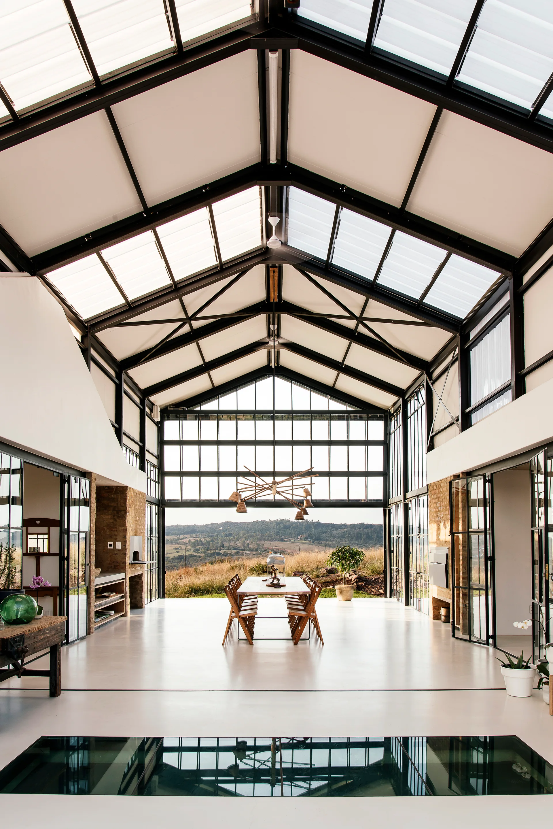 A spacious industrial dining room features a vaulted glass ceiling, steel framing, exposed brick walls, and a long wooden table with chairs.