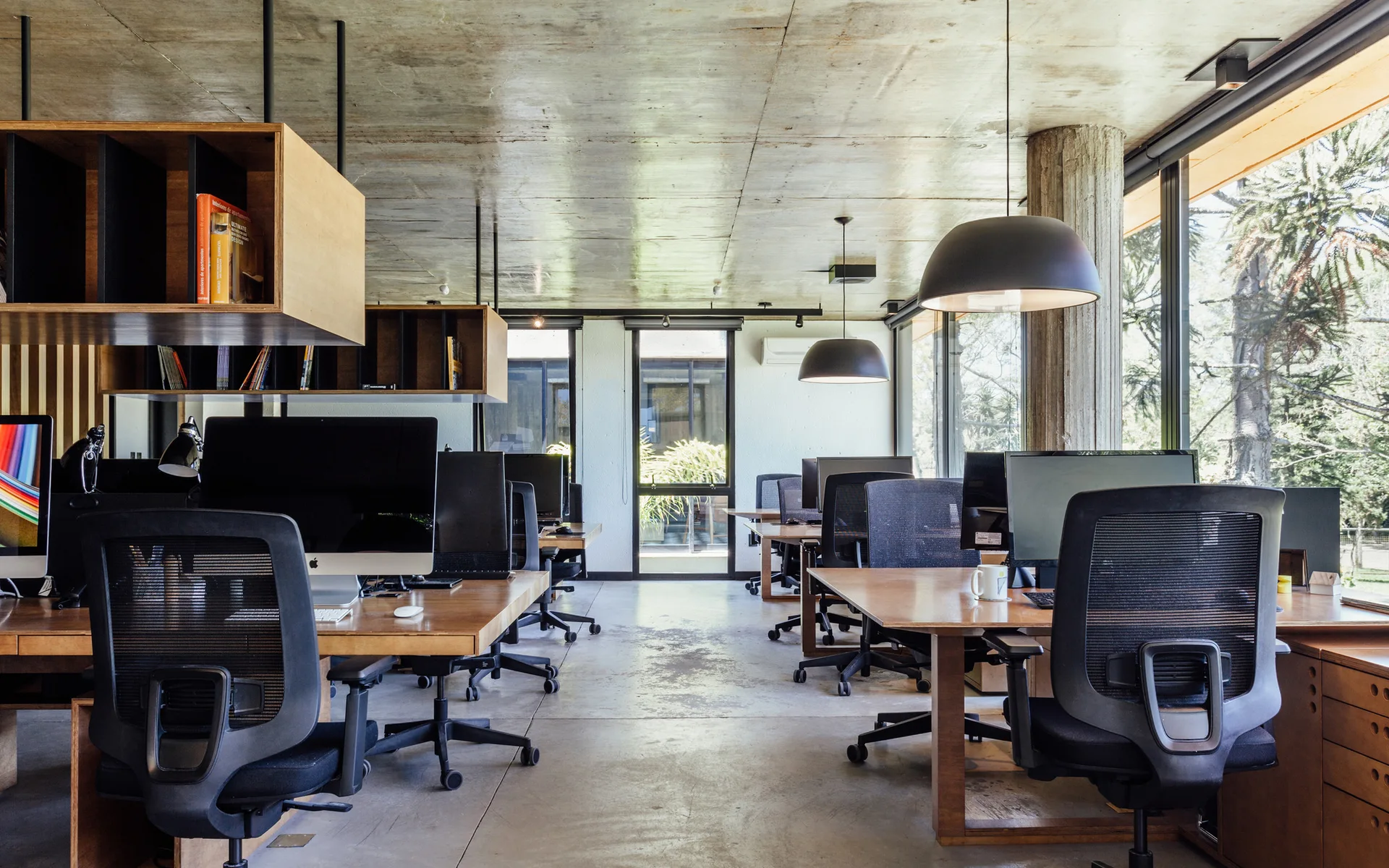 An open-plan office features wooden desks, black mesh ergonomic chairs, suspended overhead wood shelving, and an exposed concrete ceiling.