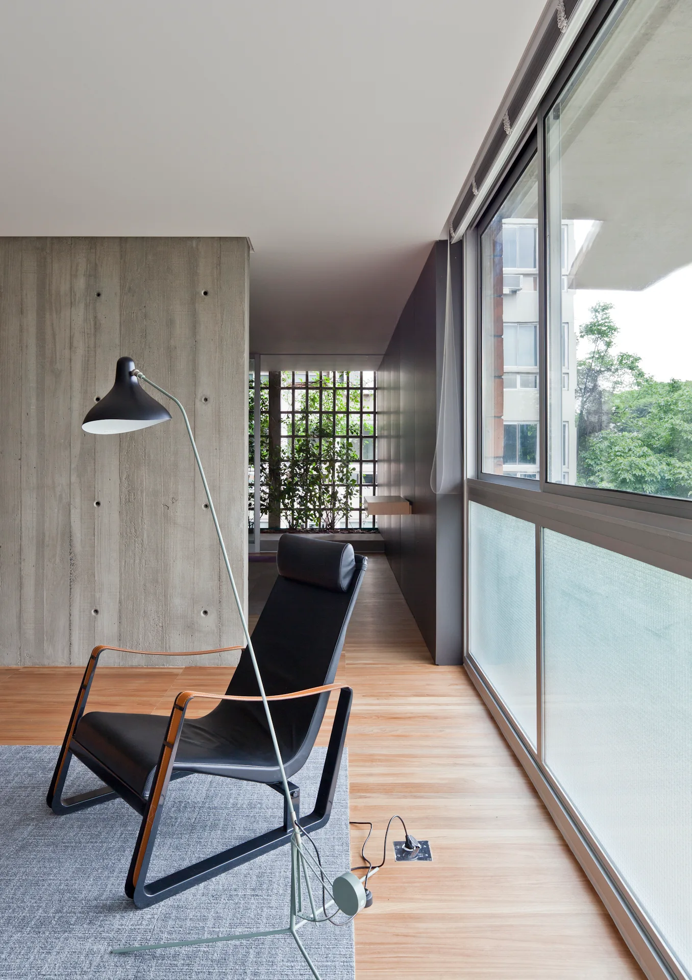 A modern minimalist living area featuring a black leather lounge chair on a gray rug next to a board-formed concrete wall and a large sliding window.