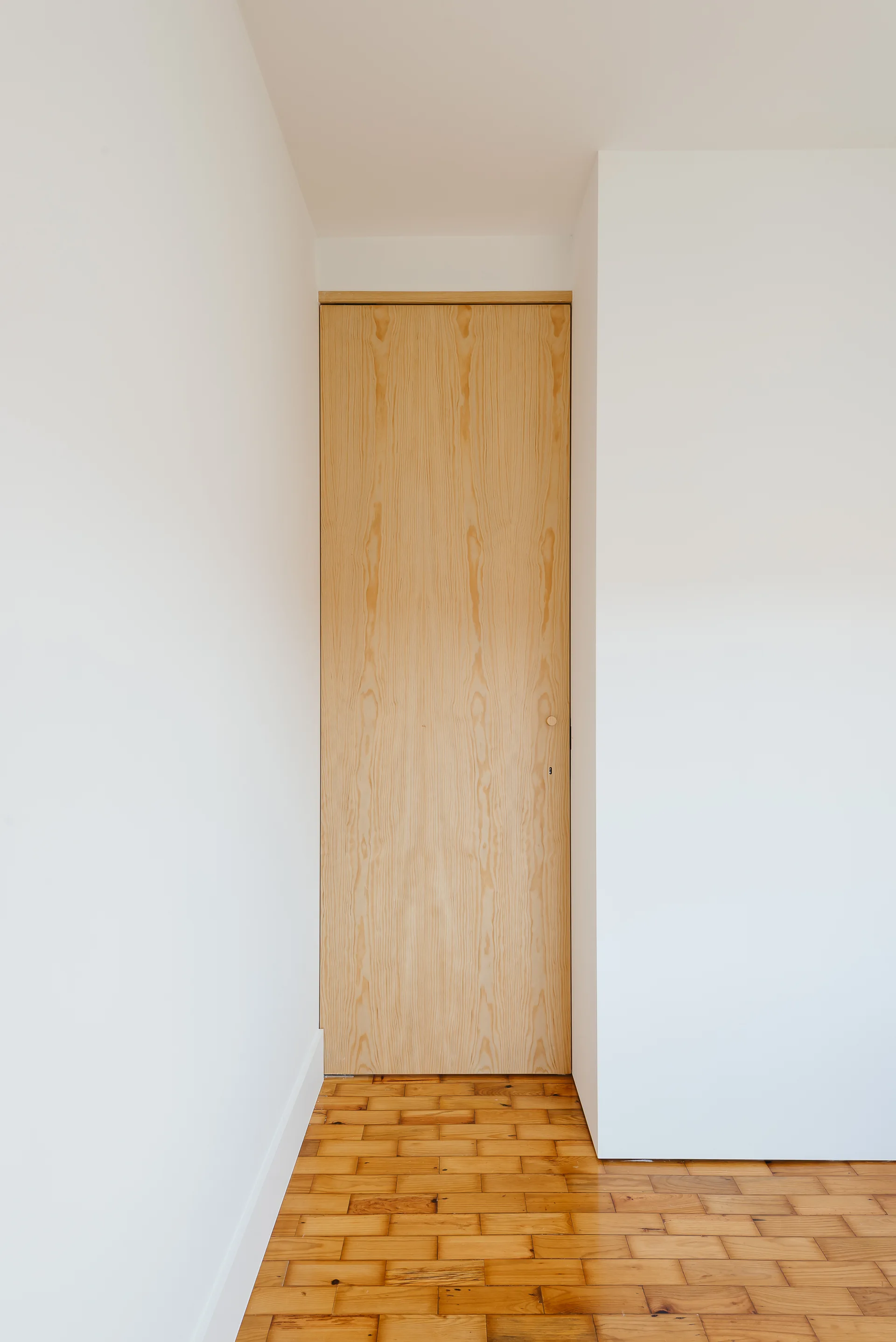 A full-height natural wood door is framed by clean white walls and warm parquet wood-block flooring in a minimalist interior.