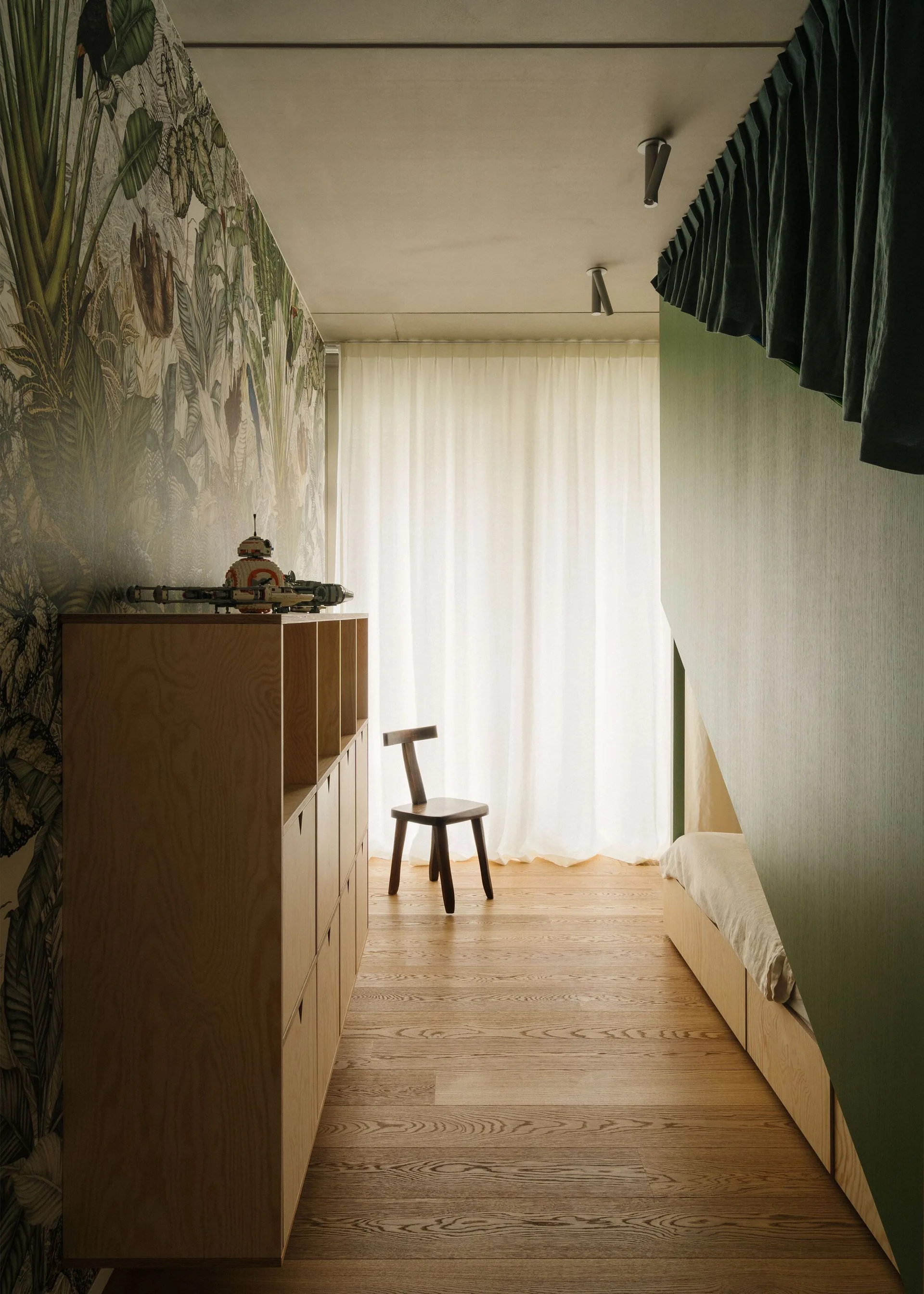 A narrow children's bedroom featuring a botanical mural wall, custom light wood storage cabinets, and a low built-in bed beneath a green partition wall.