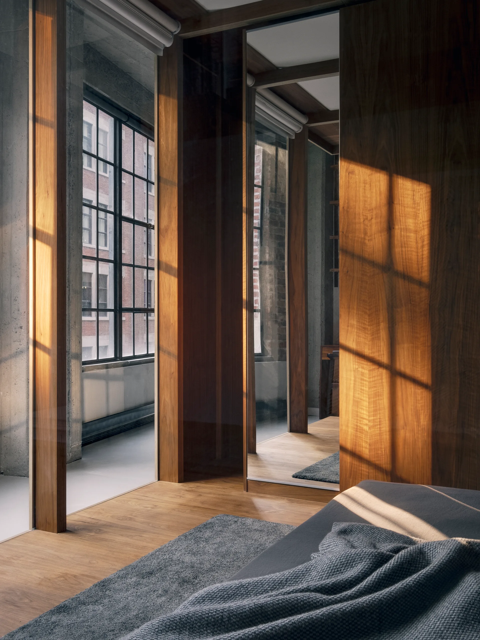 An industrial loft bedroom features warm wood-paneled walls, a mirrored wardrobe door, a grey bed throw, and a large black-framed window.