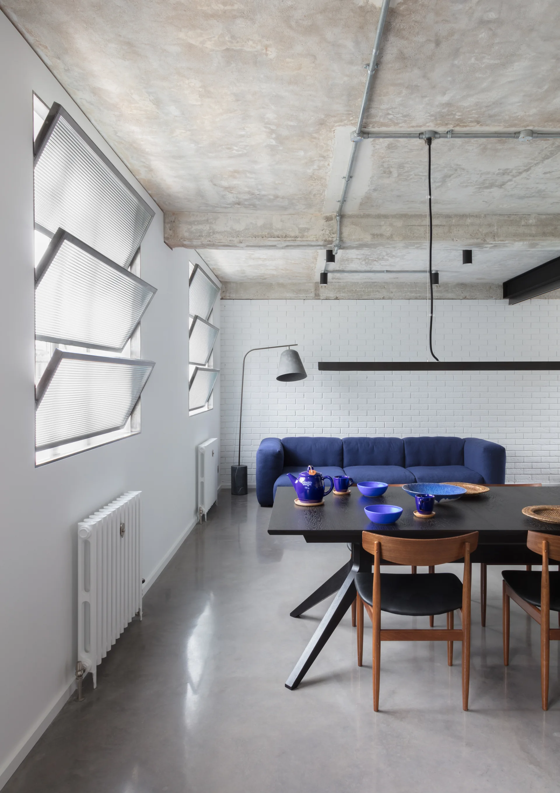 An industrial dining area with a black table, wooden chairs, polished concrete floor, and a blue sofa against a painted white brick wall.