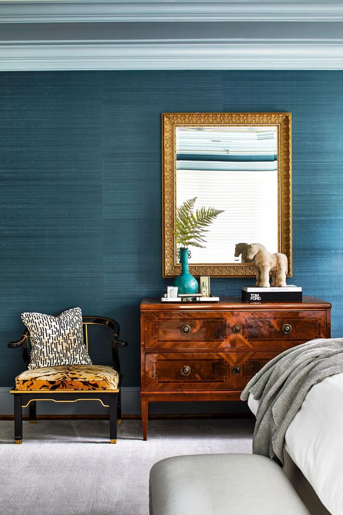A polished wood dresser topped with a gilded mirror stands against a textured blue grasscloth wall next to a black-framed accent chair.