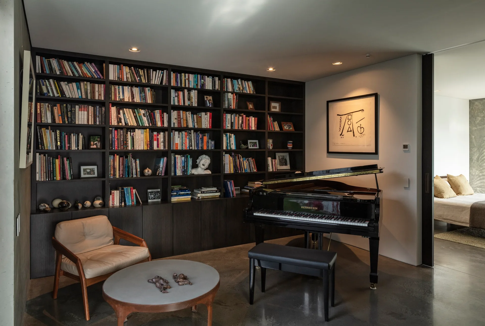 A modern home library features a dark wood floor-to-ceiling bookshelf, a black grand piano, and a leather armchair on polished concrete flooring.