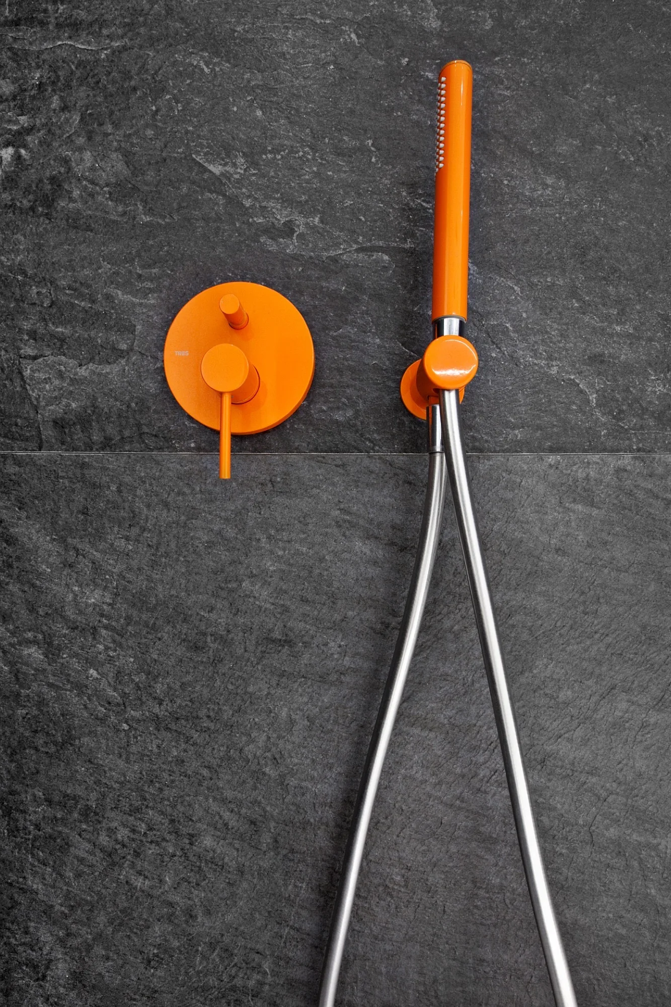 A matte orange shower control valve and handheld showerhead are mounted on a dark grey, textured slate-like wall tile.